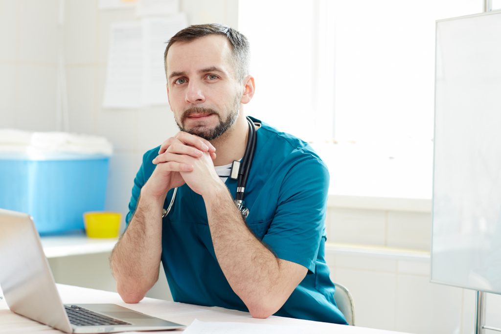 Young serious doctor in uniform looking at camera while sitting by workplace in front of laptop