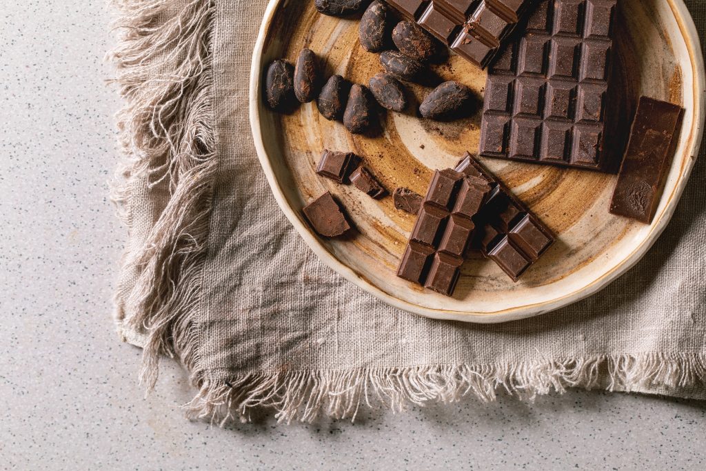 Dark and milk chocolate bar whole and chopped in ceramic plate with cocoa beans over grey linen cloth as background. Flat lay, space
