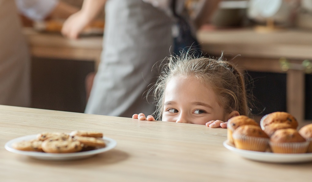 Playful little girl looking with joy at sweets, kitchen interior.
