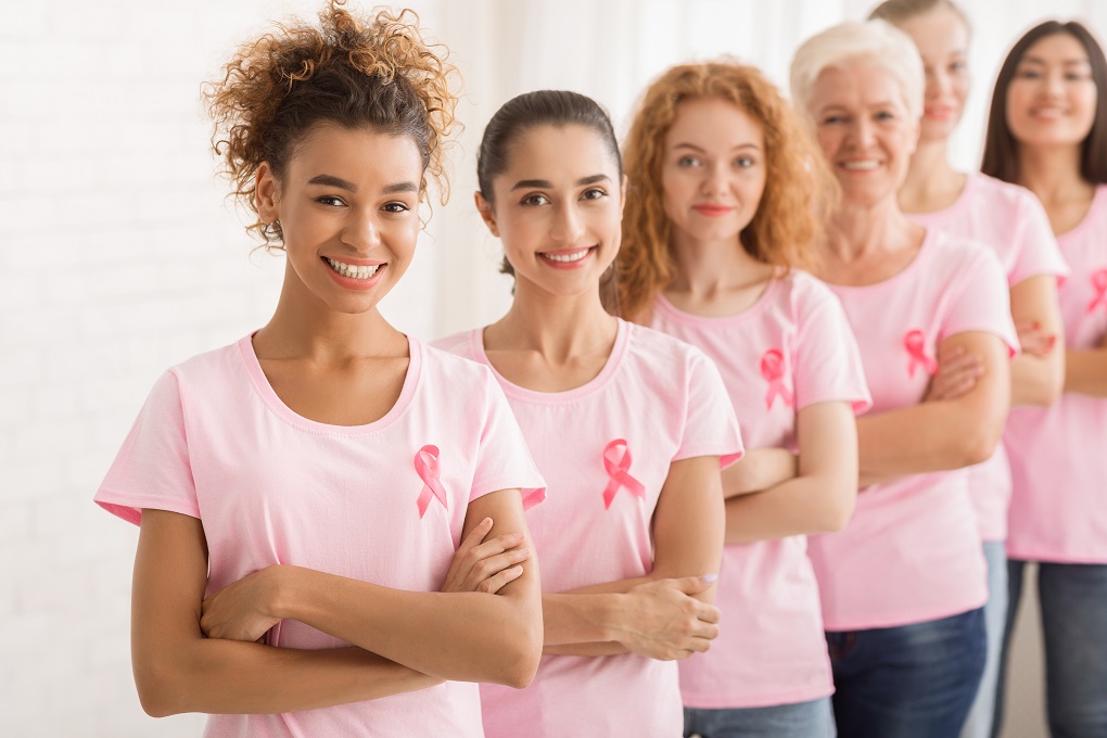 Diverse Female Volunteers In Breast Cancer Pink T-Shirts Standing In Line With Crossed Hands Over White Wall. Shallow Depth